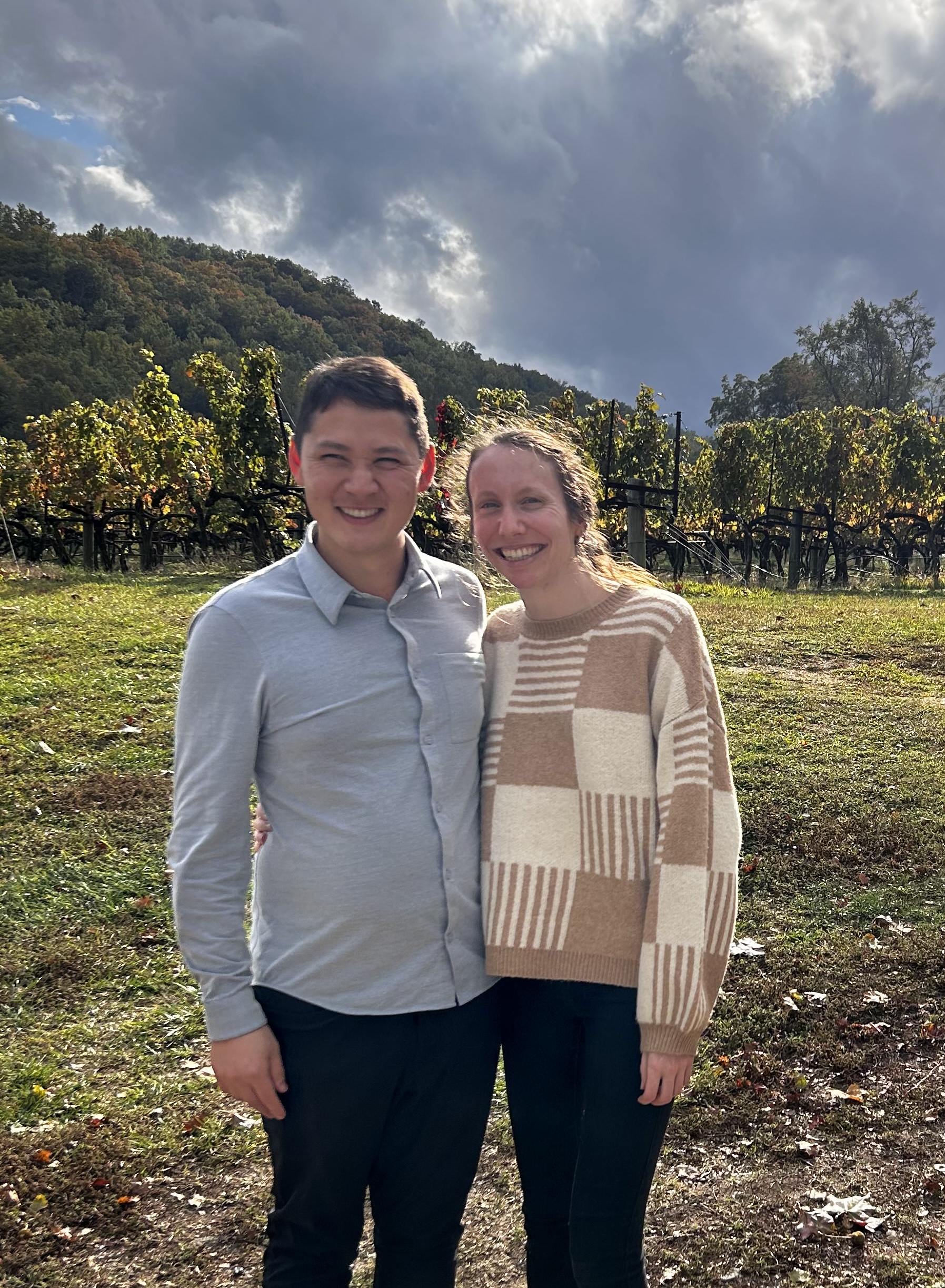 Matt and Mary smiling in a vineyard with mountains in the background.